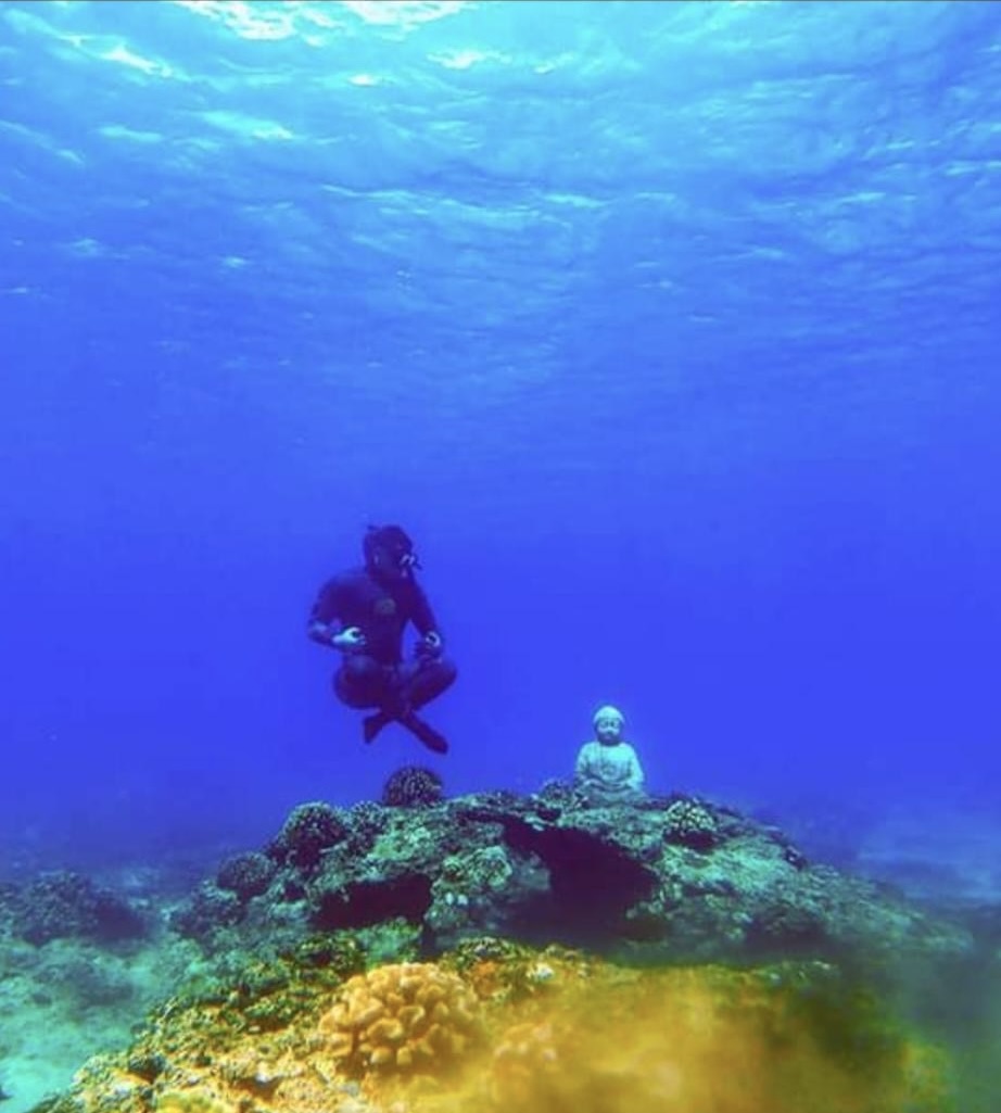 Austin Strickland freediving in lotus pose underwater next to a Buddha statue on a coral reef